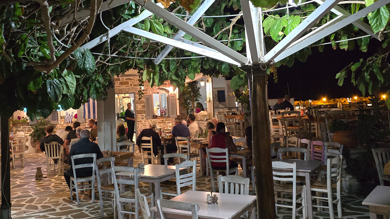 Terrasse de Kostaras sous une pergola de vigne le soir, tables blanches et ambiance de village grec, Aegiali, Amorgos