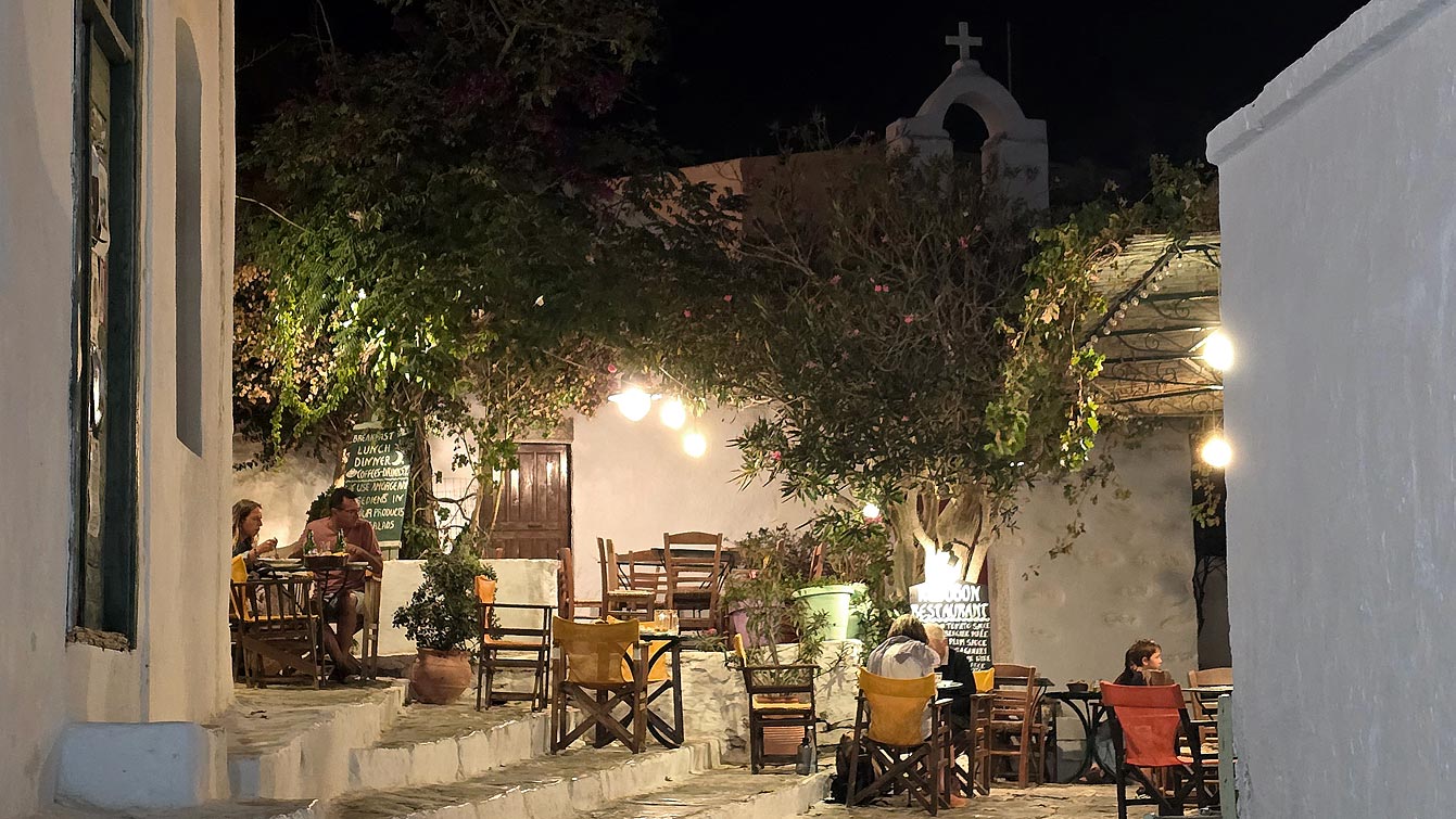 Terrasse de Kathodon sur une petite place de la Chora la nuit, tables sous les arbres et guirlandes, Amorgos
