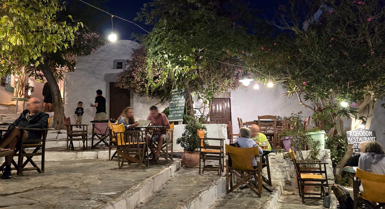 Terrasse de Kathodon sur une petite place de la Chora la nuit, tables sous les arbres et guirlandes, Amorgos