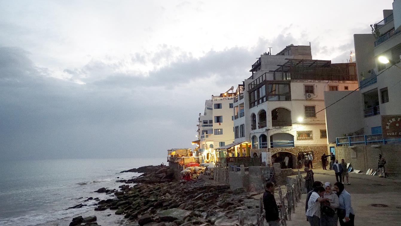 Promenade côtière de Taghazout au crépuscule, passants le long des maisons et de l'océan.