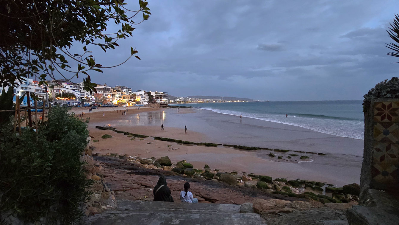 Plage de Taghazout à la nuit tombée vue depuis une terrasse rocheuse, lumières du village au fond.