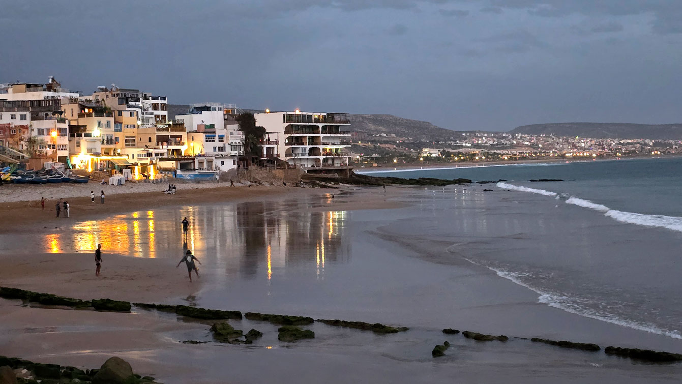 Jeunes jouant au football sur la plage de Taghazout au coucher du soleil, maisons éclairées à droite.
