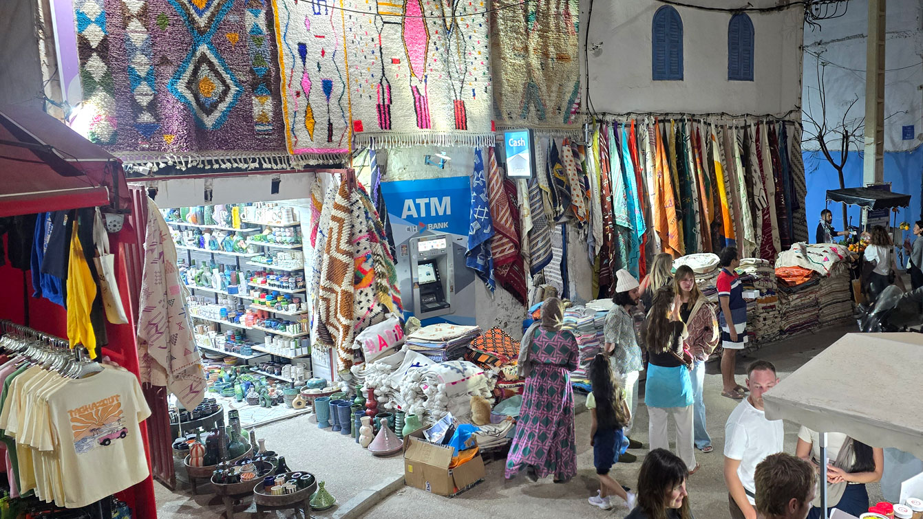 Étal de tapis et textiles berbères le long de la route à Taghazout, distributeur automatique et visiteurs le soir.