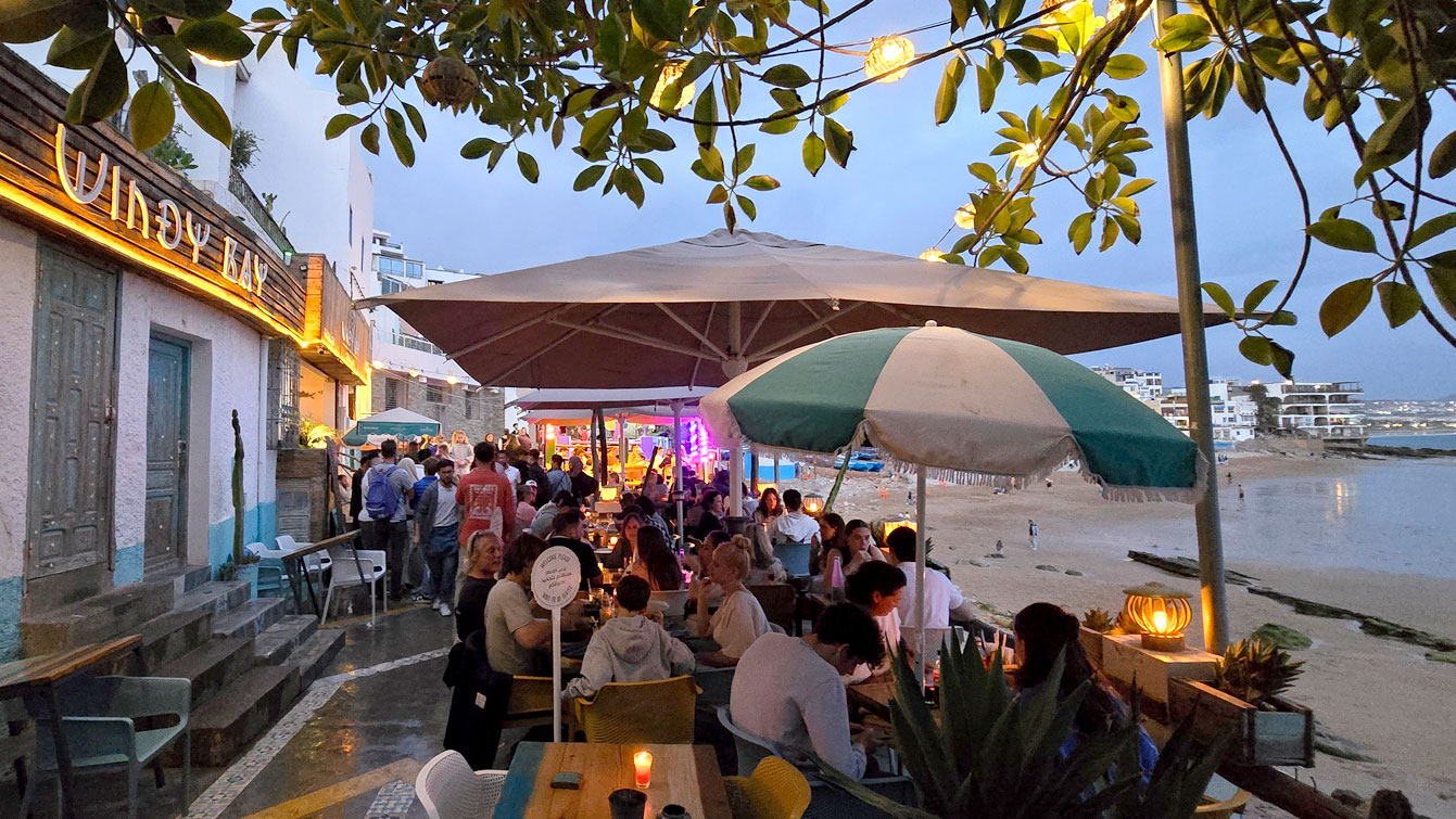 Terrasse animée du restaurant Windy Bay à Taghazout la nuit, parasols et guirlandes face à la plage.