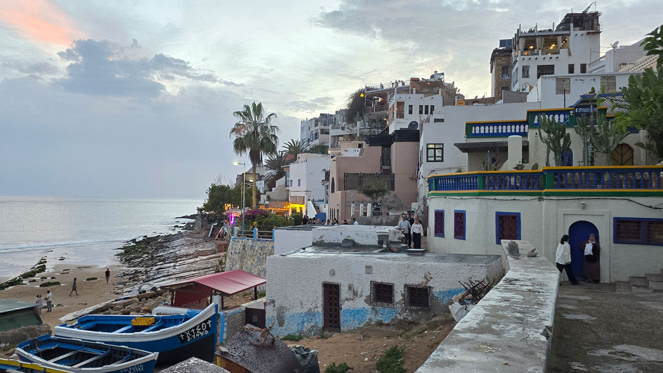 Rue côtière de Taghazout bordée de maisons et de terrasses colorées au coucher du soleil.