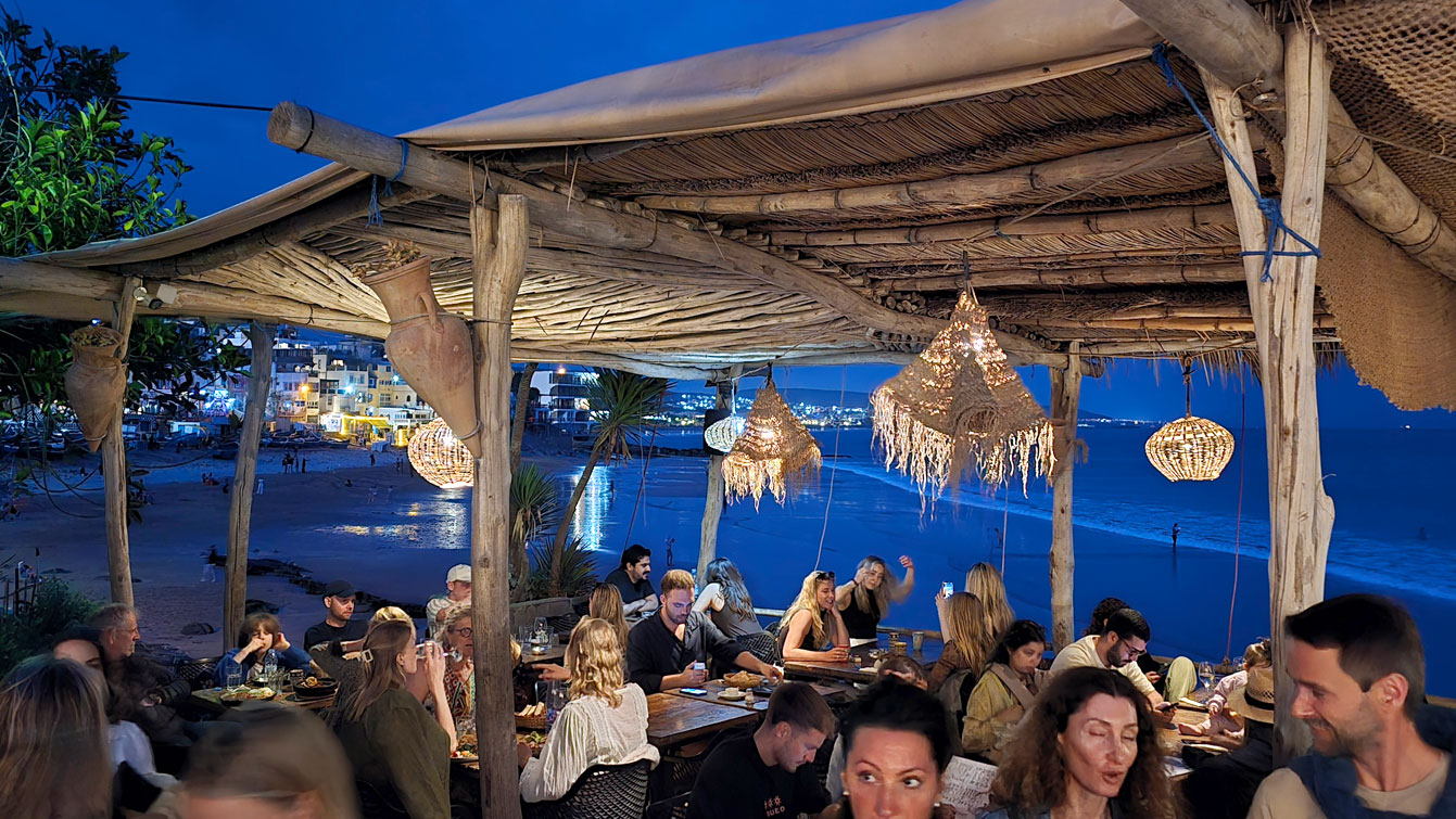 Terrasse de restaurant en bord de mer à Taghazout la nuit, paillotes en chaume et guirlandes lumineuses.