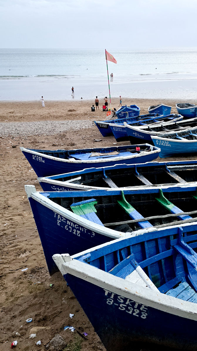 Barques de pêche bleues alignées sur la plage de sable de Taghazout, drapeau et baigneurs au loin.